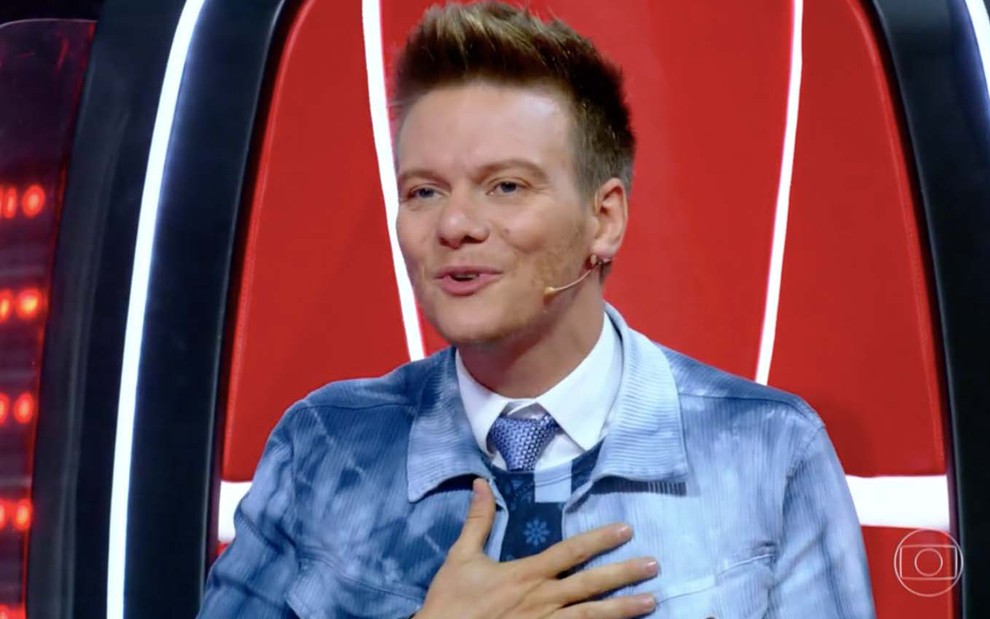 O cantor Michel Teló leva a mão ao peito sentado na cadeira vermelha do The Voice Kids