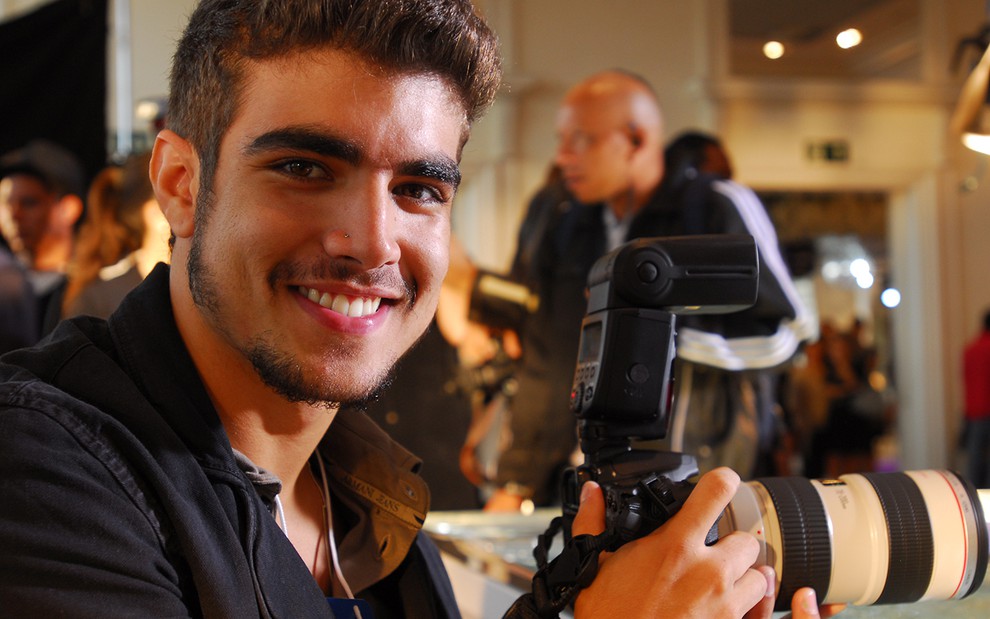 O ator Caio Castro caracterizado como Edgar, ele segura uma câmera fotográfica profissional diante de uma passarela de desfiles em cena de Ti Ti TI
