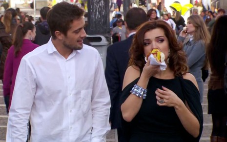 Rafael Cardoso e Mayana Neiva em cena de Ti Ti Ti: atores estão na rua e atriz segura cachorro quente com a mão direita