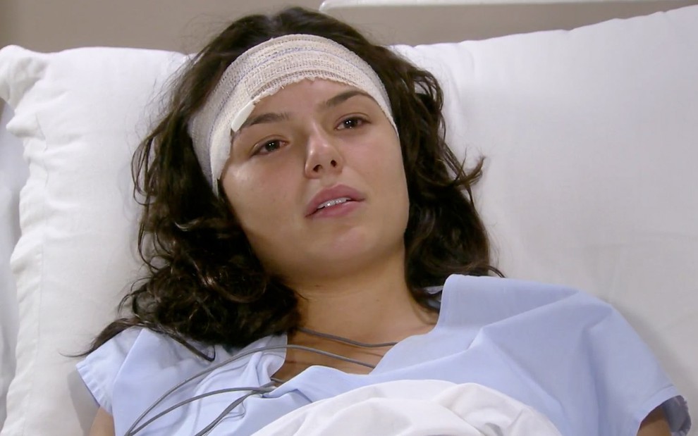 Isis Valverde em cena de Ti Ti Ti: deitada em cama hospital, atriz está com a cabeça enfaixada e usa roupa de paciente