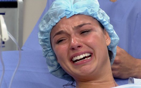 Isis Valverde em cena de Ti Ti Ti: caracterizada como Marcela, atriz está em maca de hospital, roupa cirúrgica e olha com emoção para algo fora do quadro