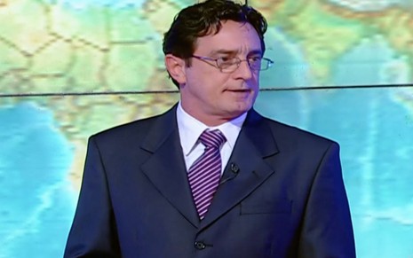 O ator Eduardo Galvão a frente de um mapa-mundi como o apresentador Douglas Sábato em cena de Toma Lá Dá Cá