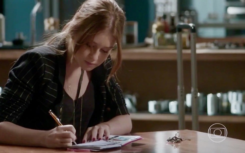 Sentada em uma mesa, a atriz Marina Ruy Barbosa escreve em caderno em cena da novela Totalmente Demais