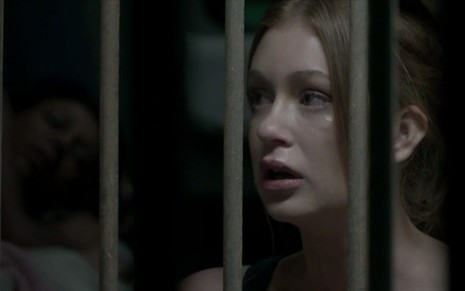 Com cara de assustada, Marina Ruy Barbosa coloca o rosto na grade de uma cela em cena da novela Totalmente Demais