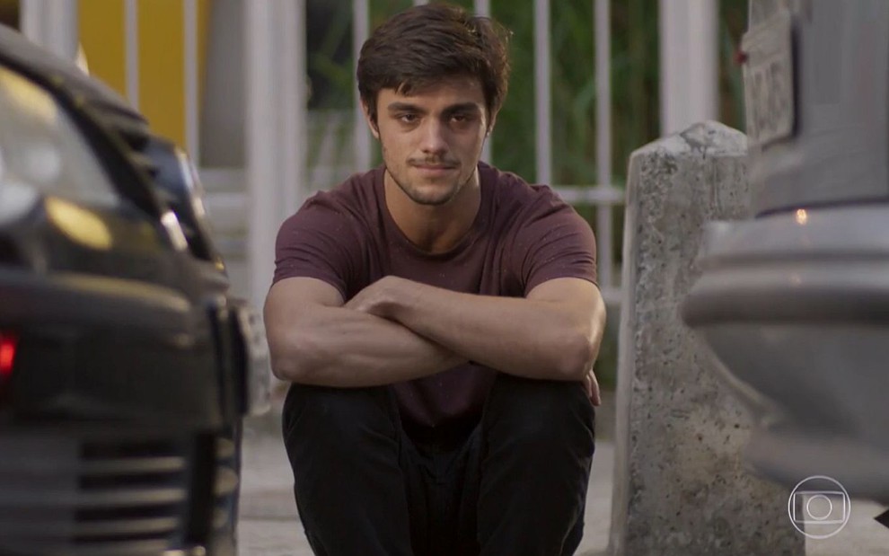 Jonatas (Felipe Simas) sentado na guia da calçada, triste, em cena de Totalmente Demais