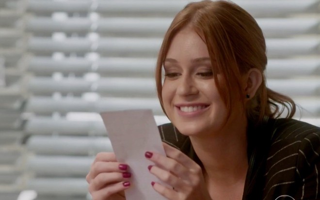A atriz Marina Ruy Barbosa segura um papel diante do rosto e sorri em cena como Eliza em Totalmente Demais