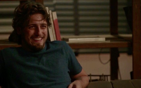 O ator Daniel Rocha, sentado em um sofá, sorri de maneira sarcástica durante gravação da novela Totalmente Demais