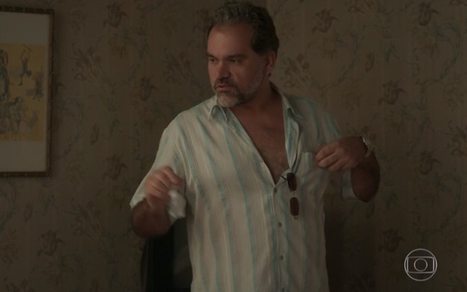 O ator Rodrigo Rangel, com uma camisa de botões aberta no peita, um óculos escuro pendurado no bolso da blusa, em cena como delegado Peçanha na novela Totalmente Demais