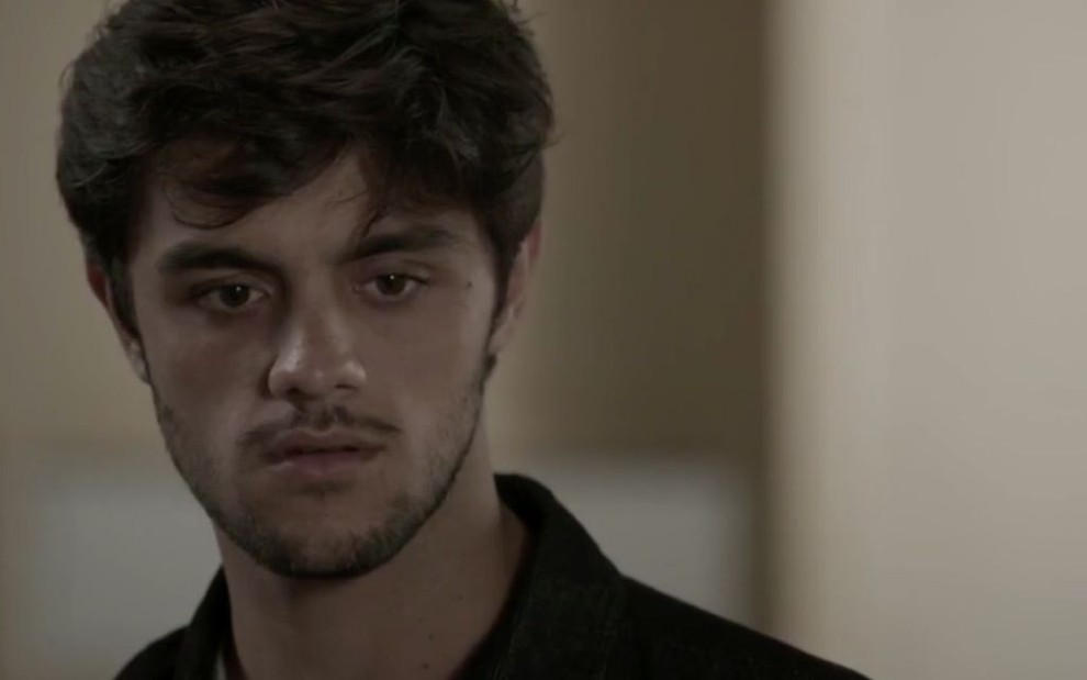 O ator Felipe Simas, com a expressão preocupada, usando uma camiseta polo preta, em cena como Jonatas na novela Totalmente Demais