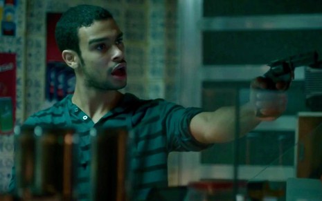 O ator Sergio Malheiros em cena de Totalmente Demais: personagem usa camiseta marrom e aponta arma para alguém fora do quadro