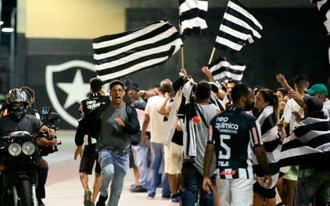 O ator Cauã Reymond corre caracterizado como Christian enquanto torcedores do Botafogo agitam bandeiras ao fundo em gravação de Um Lugar ao Sol