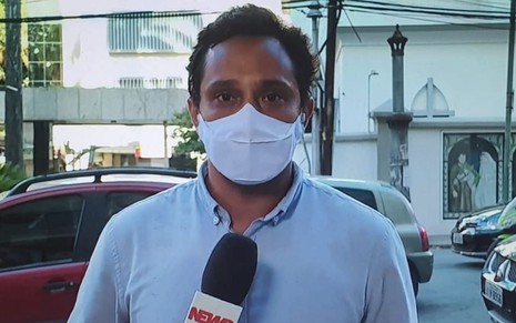 O repórter Pedro Neville com um microfone na mão direita e máscara em frente a um hospital