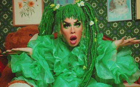 Gloria Groove com uma fantasia verde e cara de assustada em cima de uma cama