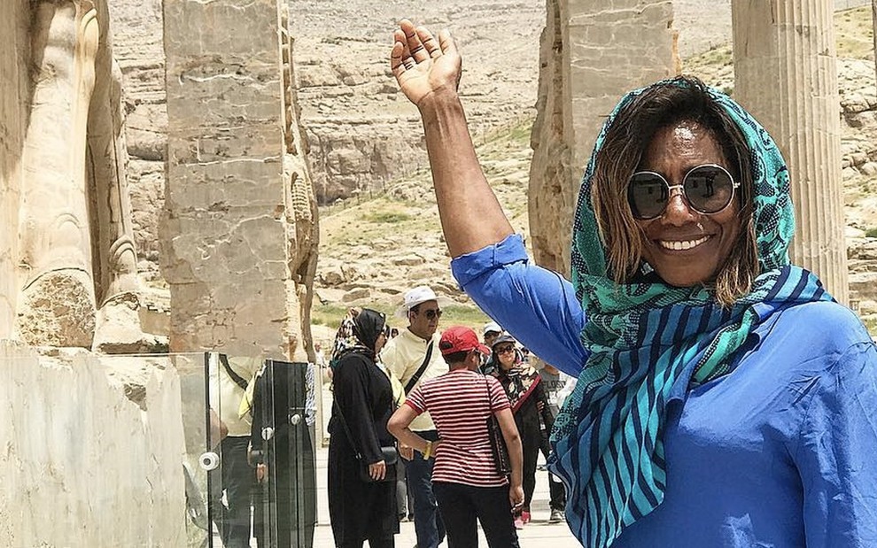 A jornalista Gloria Maria com um véu ergue a mão e aponta uma ruína em Persépolis, no Irã