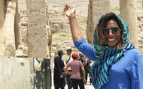 A jornalista Gloria Maria com um véu ergue a mão e aponta uma ruína em Persépolis, no Irã