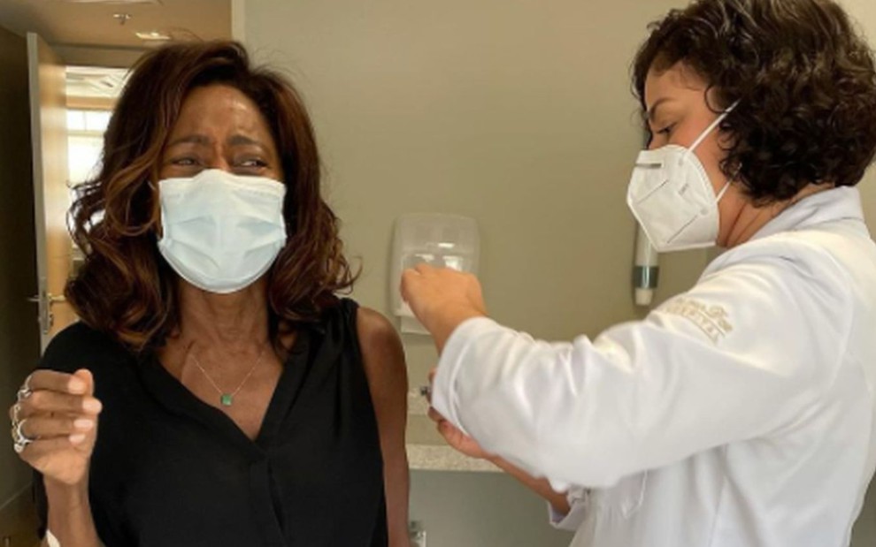 Gloria Maria de máscara branca, roupa preta, tomando a vacina no braço esquerdo, aplicada pela enfermeira, de jaleco branco