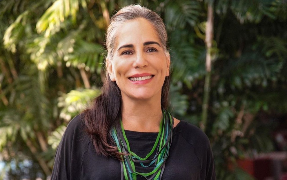 Gloria Pires está em frente a um local cheio de árvores, ela usa camiseta preta e colar verde; ela sorri e está com o cabelo preso