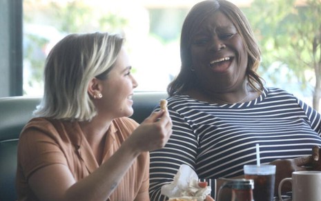 Em um restaurante, Mae Whitman segura nos dedos uma batata frita enquanto ri junto a Retta, sentada ao seu lado em cena de Good Girls
