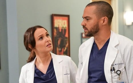 Perto da recepção de um hospital, Camilla Luddington olha para Jesse WIlliams; ambos vestem jaleco de médicos em Grey's Anatomy
