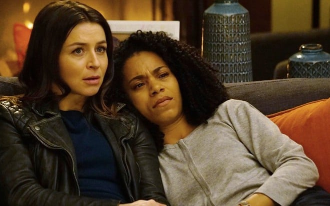 Esparrramada em um sofá, Kelly McCreary encosta a cabeça no ombro esquerdo de Caterina Scorsone em cena de Grey's Anatomy