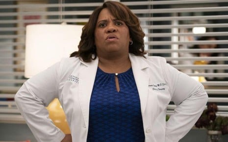 A atriz Chandra Wilson, com um jaleco de médica, coloca as mãos na cintura e faz cara de frustrada em Grey's Anatomy