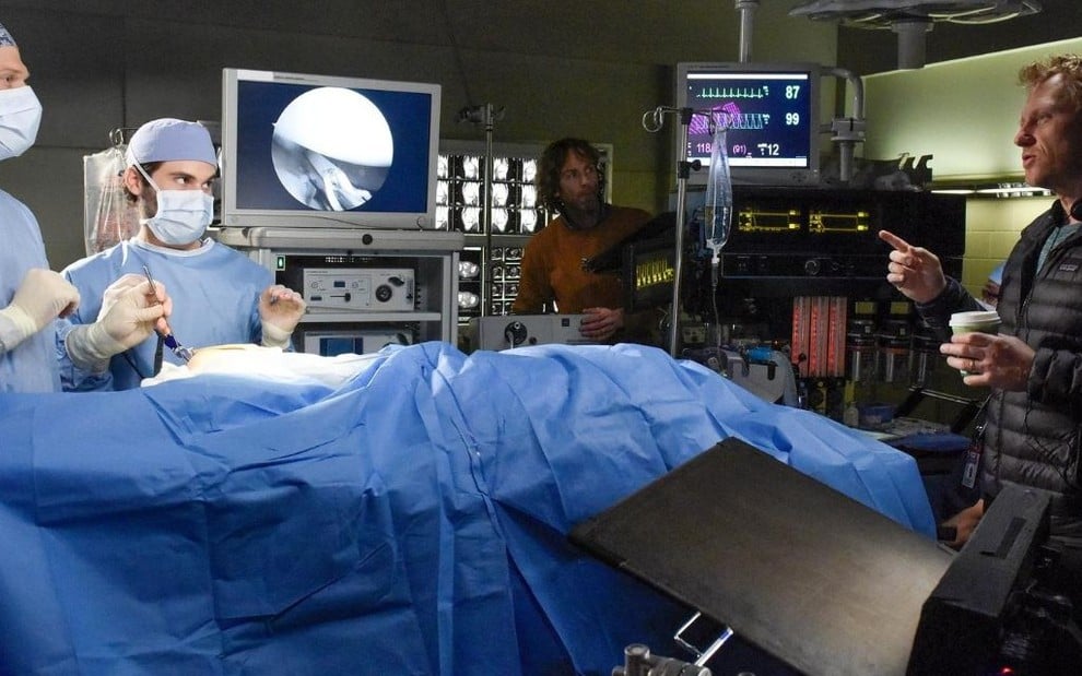 Dentro de uma sala de cirurgia, os atores Chris Carmack e Jake Borelli ouvem o diretor Kevin McKidd em Grey's Anatomy