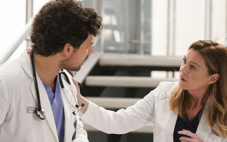 Com um estetoscópio no pescoço, Giacomo Gianniotti olha para Ellen Pompeo, que toca o seu ombro esquerdo em Grey's Anatomy