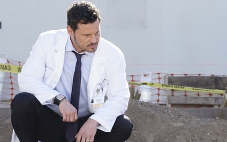 Agachado e vestindo um jaleco de médico, Justin Chambers olha triste para uma cova em episódio de Grey's Anatomy