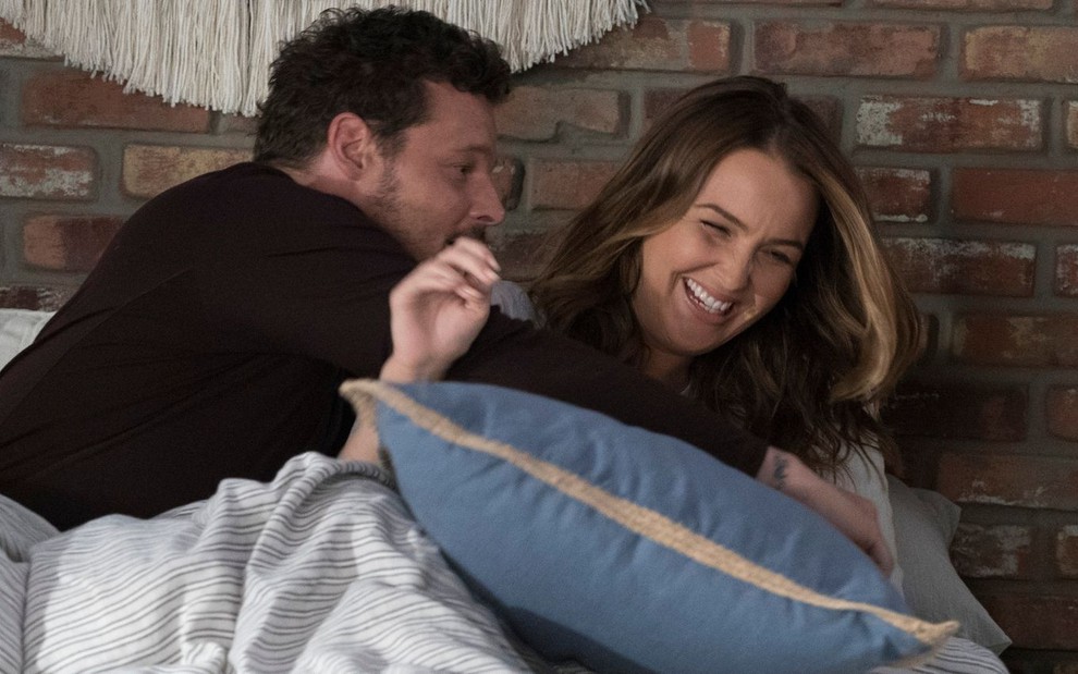 Deitados em uma cama, Justin Chambers e Camilla Luddington brincam de se agarrar em cena de Grey's Anatomy