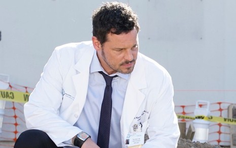 Com um jaleco de médico, Justin Chambers aparece agachado na beira de uma vala em episódio de Grey's Anatomy