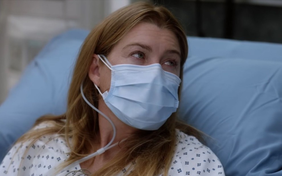 Ellen Pompeo, intérprete de Meredith Grey, de máscara deitada na cama do hospital olhando para o lado