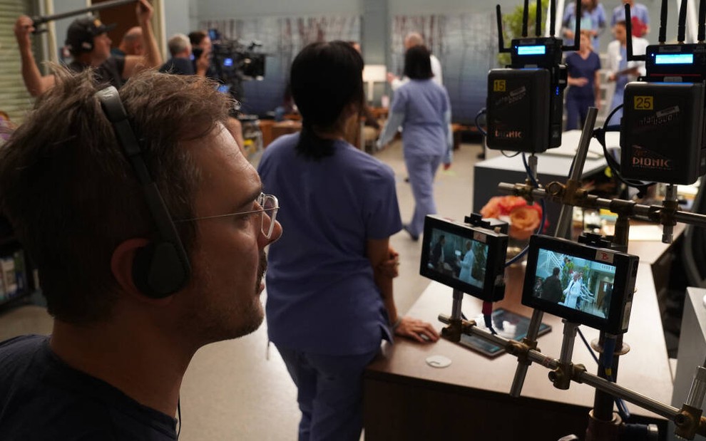 De óculos, o diretor Michael Medico olha para pequenos monitores enquanto uma cena de Grey's Anatomy é gravada
