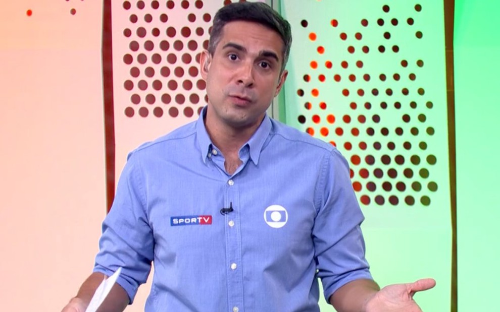 Gustavo Villani com camisa azul e braços abertos no cenário de um estúdio da Globo