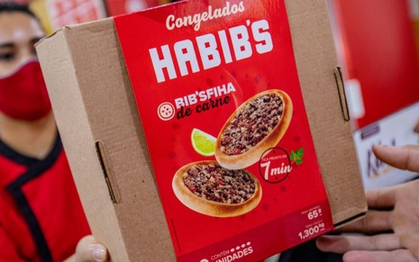 Imagem de funcionária não identificável na foto entregando caixa de esfirras congeladas do Habib's para cliente