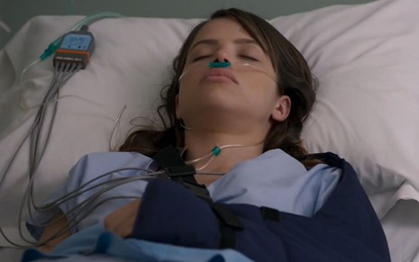 A atriz Agatha Moreira aparece deitada em uma maca de hospital, com os olhos fechados e oxigênio no nariz em cena como Camila de Haja Coração
