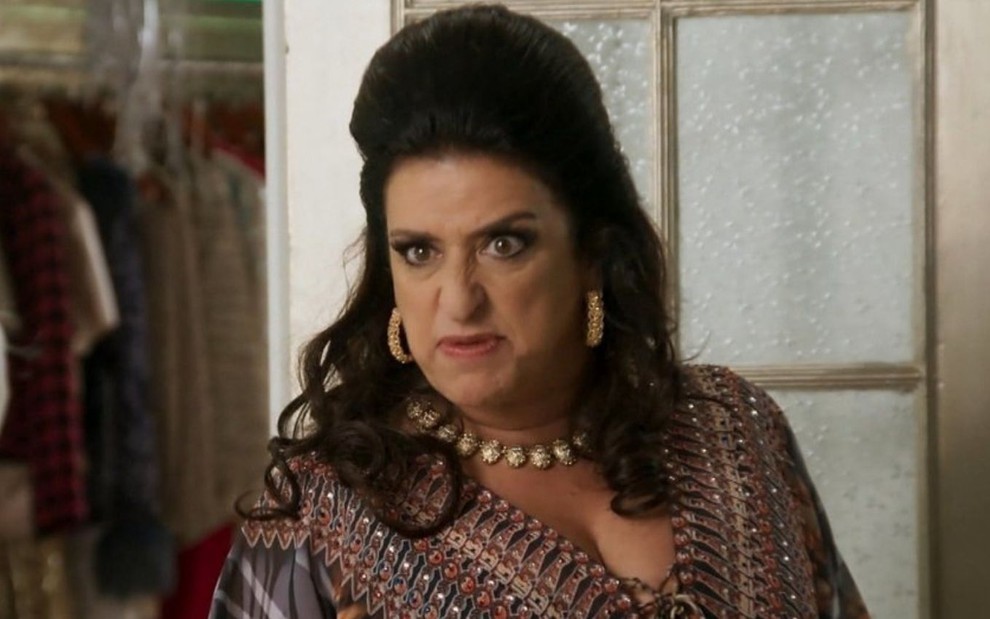 A atriz Grace Gianoukas usa um topete enorme nos cabelos e faz expressão nervosa em cena como Teodora em Haja Coração