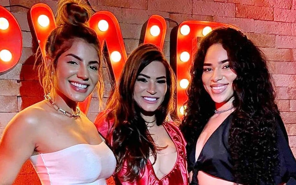 Hariany Almeida está usando um top branco, cabelo preso e aparece sorridente na foto; ao lado dela está Raissa Barbosa com um vestido rosa e cabelo solto; ela sorri para a foto; ao lado da ex-Fazenda está Elana Valenaria com um vestido preto e cabelo solto