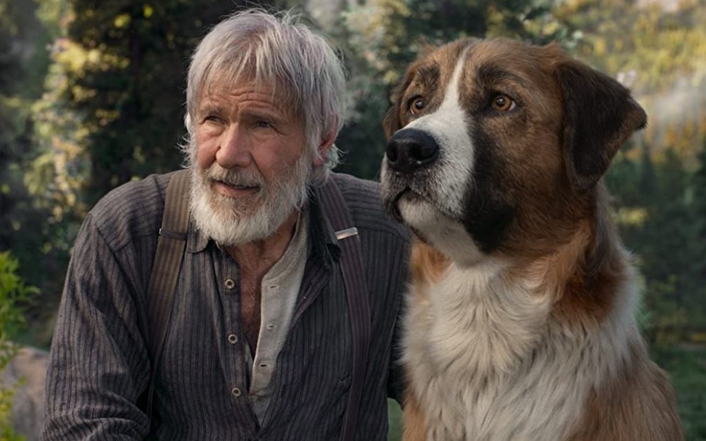 No meio da mata, o personagem John Thornton (Harrison Ford) está sentado ao lado do cão Buck em O Chamado da Floresta (2020)