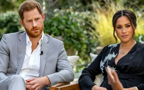 Harry ao lado de Meghan Markle sentados durante entrevista; ele está de terno claro e camisa, e ela, de vestido preto decotado
