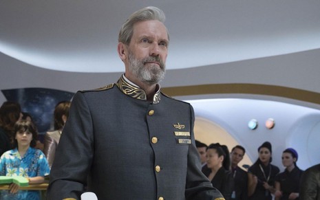 O ator Hugh Laurie dirige uma nave vestido com um casaco escuro de capitão com botões dourados