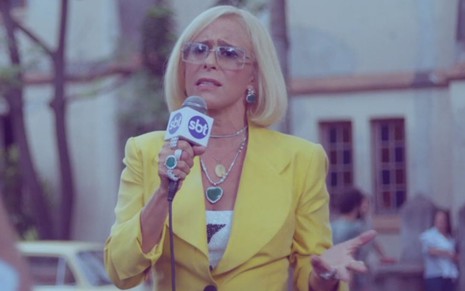 Com microfone do SBT e usando uma roupa amarela, Andréa Beltrão está como a personagem Hebe em série da Globo
