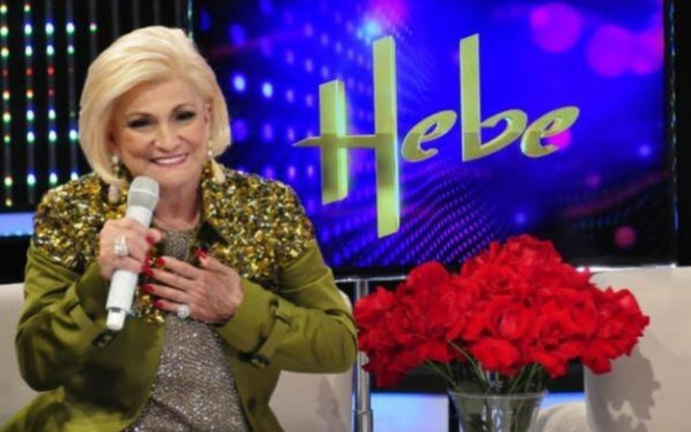 Usando casaco verde, Hebe Camargo segura microfone branco ao lado de vaso com rosas e e tela de TV escrito 'Hebe'