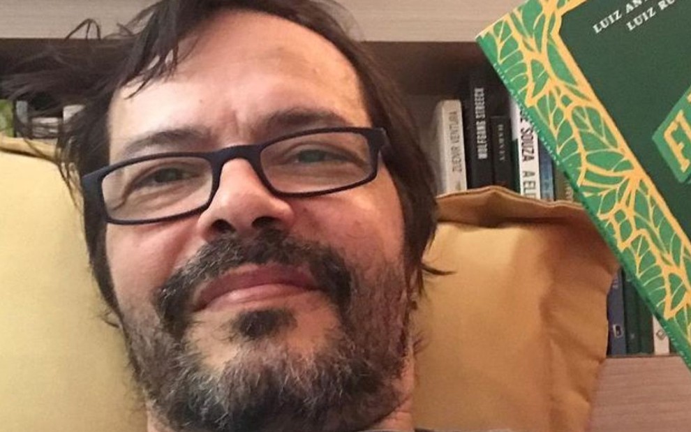 Heitor Martinez, de óculos e segurando livro, posa para foto encostado em almofada em sua casa