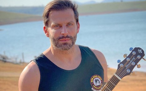 O ator Henri Castelli de regata preta, em frente a um lago, segurando um violão
