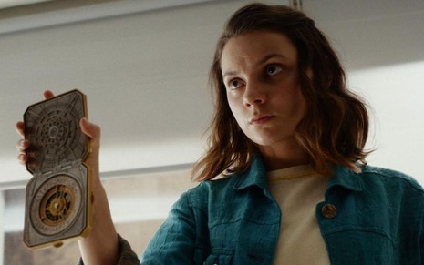 Com uma roupa verde, Dafne Keen segura um distintivo na mão direita em cena da série His Dark Materials