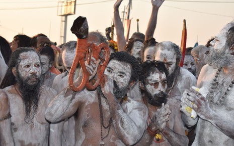 Homens pintados de branco celebram em multidão, um deles segura um instrumento de sopro que parece uma cobra
