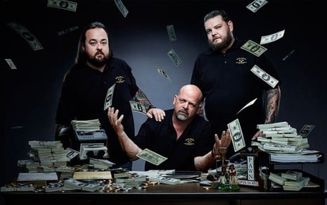 Chumlee Russell, Rick e Corey Harrison, do Trato Feito, estão diante de uma mesa cheia de dinheiro, com algumas notas no ar