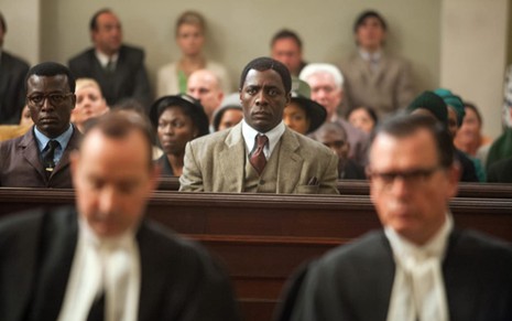 Idris Elba focado ao centro, sentado de terno e gravata com um feição séria; pessoas à sua volta, no que parece ser um tribunal