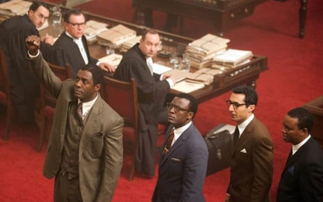 Em um tribunal Idris Elba como Mandela (à esq),  seguido por Tony Kgoroge, Riaad Moosa e Thapelo Mokoena, todos vestido com ternos e atrás três homens sentados, vestidos com beca de advogado