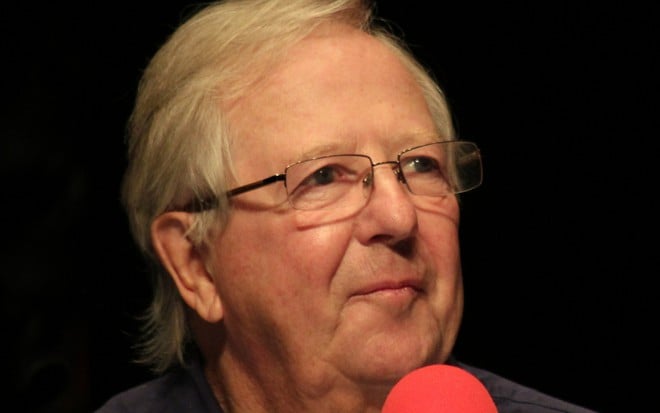 De óculos e cabelo grisalho, o comediante Tim Brooke-Taylor fala em um microfone no programa de rádio I'm Sorry I Haven't a Clue